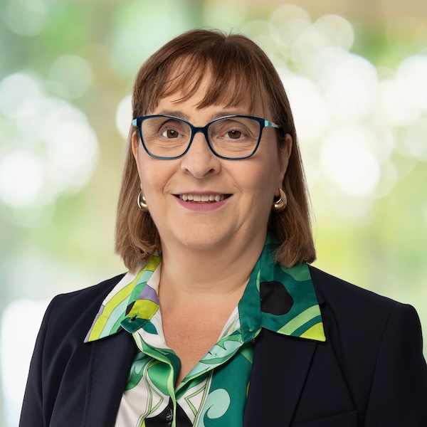 Porträt von Maria Lovecchio mit kurzem braunem Haar, blauer Brille und gemusterter grüner Bluse vor unscharfem Hintergrund