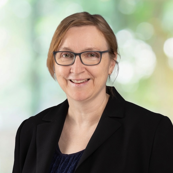 orträt von Rebecca Höss mit Brille und zusammengebundenem Haar in schwarzem Blazer vor hellem, verschwommenem Hintergrund