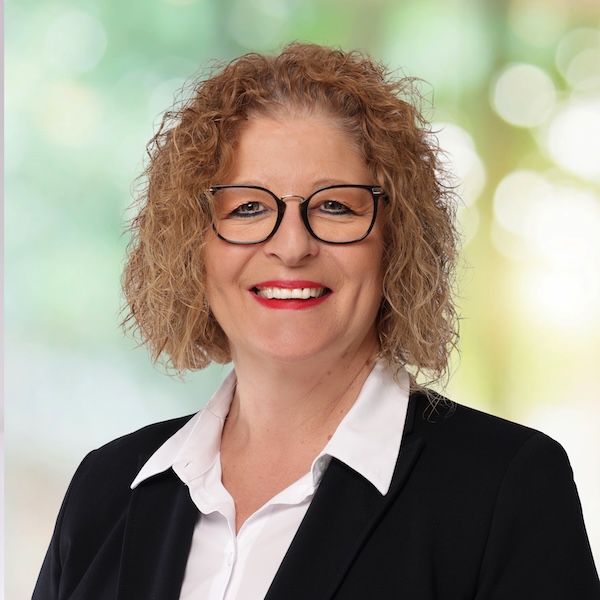 Porträt von Sandra Ferrante mit lockigem, schulterlangem Haar, Brille und rotem Lippenstift vor hellem Hintergrund