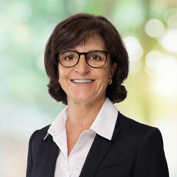 Porträt von Silvia Wenger mit Brille und dunklem Blazer vor unscharfem grünem Hintergrund