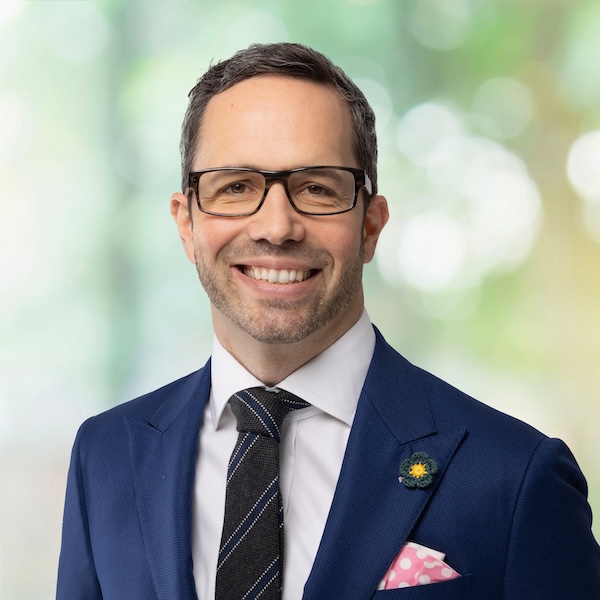 Porträt von Simon Dietrich im Anzug mit Brille und Einstecktuch, vor unscharfem grünem Hintergrund